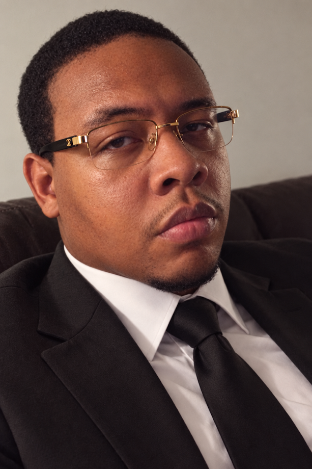 Larry Tha Volkano Stamps Jr. wearing glasses, a black suit, white shirt, and black tie